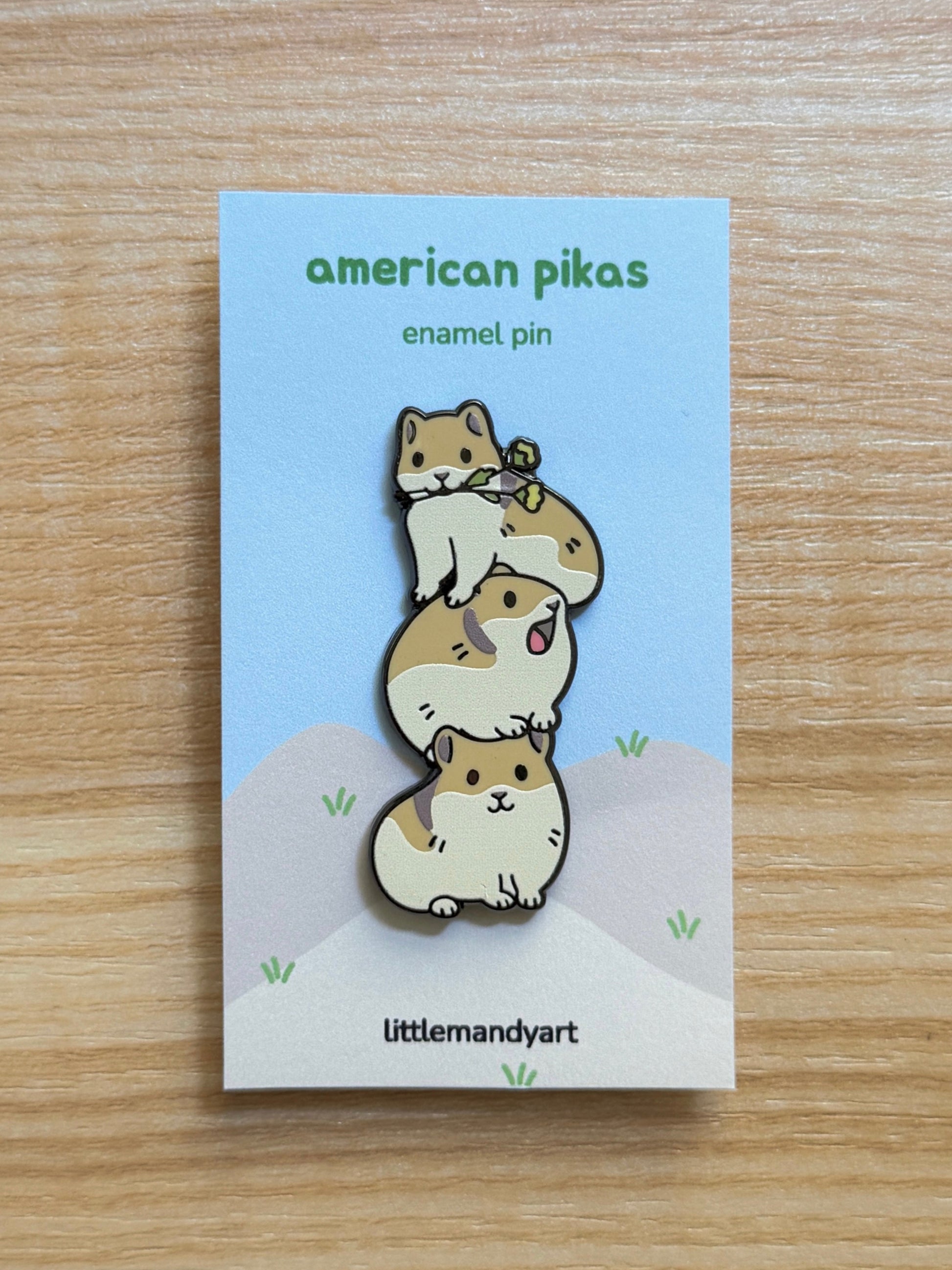 Enamel pin of American pikas on a card with 'littlemandyart' branding, placed on a wooden surface.